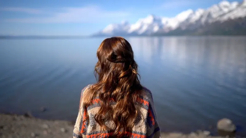 A woman looks out across a calm lake toward snow-capped mountains